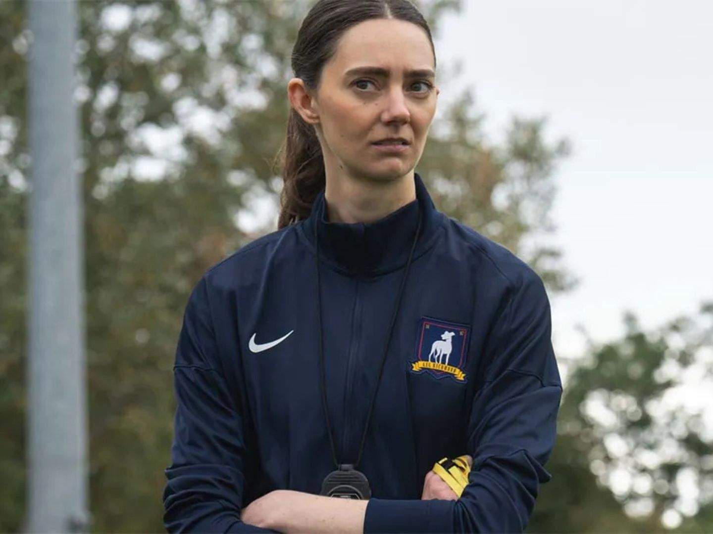 Coach Alice Chilton (Tanya Reynolds) in Ted Lasso season 4 episode 2 watching her team (not pictured) train with her arms crossed and a stopwatch hanging over her neck while holding a whistle.
