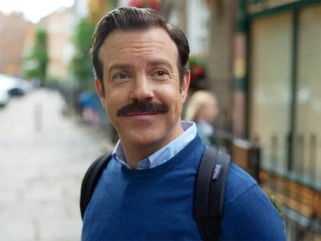 Ted Lasso (Jason Sudeikis) steps outside into a London street and looks at the view happily yet nostalgically as he begins to walk wearing a backpack and a dark blue sweater.