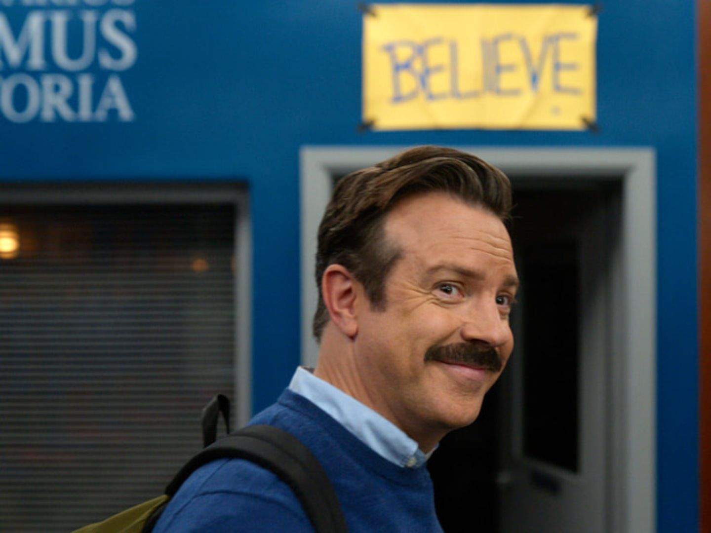 Ted Lasso (Jason Sudeikis) proudly and nostalgically walks around the locker room of the men's team he formerly coached years ago. He is wearing a backpack and dark blue sweater. The iconic "Believe" sign that Lasso put up above the door to remind his team about belief can be seen in the photo.