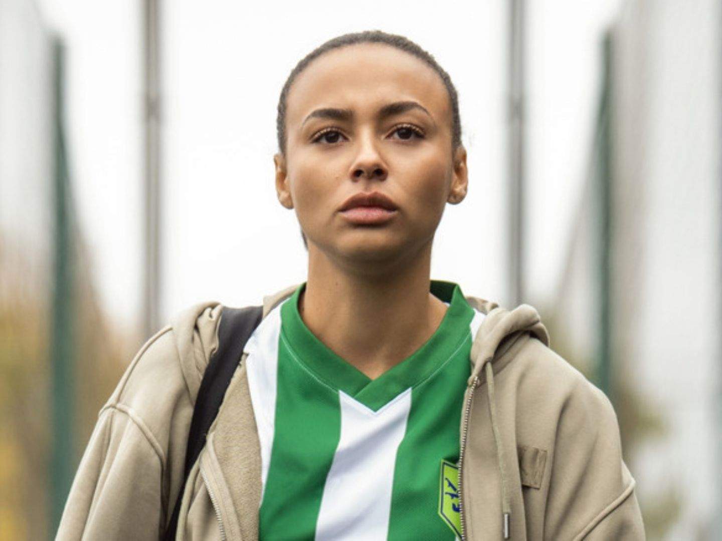 Ted Lasso season 4 newcomer Gemma (Abbie Hern) makes her first appearance in a green and white football jersey and jacket, wearing a bag with one strap on her right shoulder, leaving a football pitch.