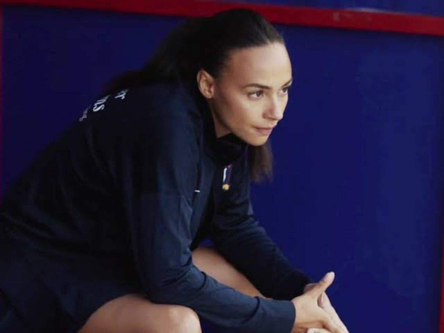 Ted Lasso season 4 newcomer Gemma (Abbie Hern) is ready to play. Ponytailed and wearing the team jacket and shorts, she is sitting down waiting to be called onto the field.