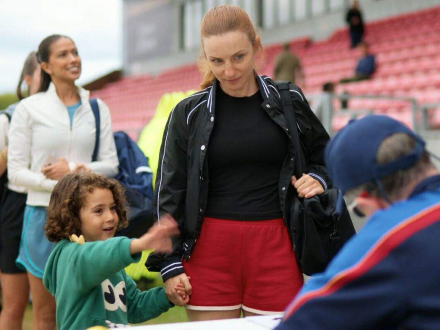 Ted Lasso season 4 newcomer Lizzie Davis (Faye Marsay)  signs up for tryouts , bringing her toddler child with her because the decision to tryout was spontaneous and she couldn't find childcare immediately. Other candidates stand behind her in a line.