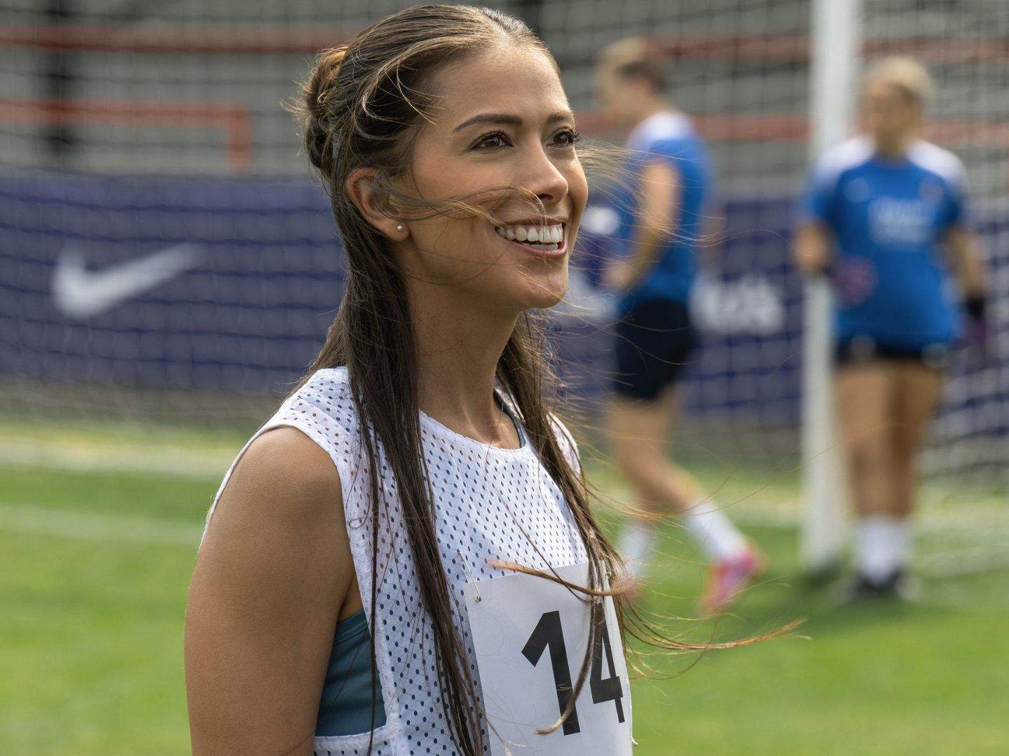 Ted Lasso season 4 guest Maria Rojas (Paloma Cinco) wearing a sleeveless shirt smiling with a goalpost in the background. Two football trainees are also in the background.
