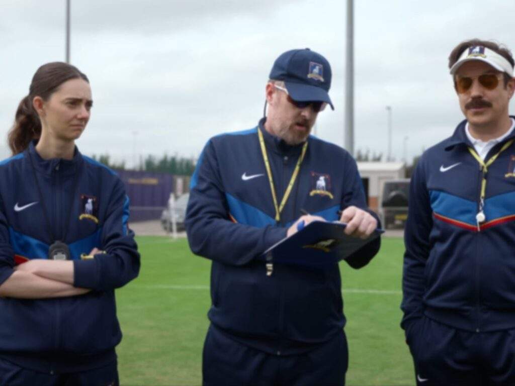 From right to left: Ted Lasso (Jason Sudeikis), Coach Beard (Brendan Hunt), and Alice Chilton (Tanya Reynolds) assess the women's football team tryouts. Beard holds a clipboard as Ted (in sunglasses) and Alice look at the candidates. All are wearing uniformed training attire (jacket).