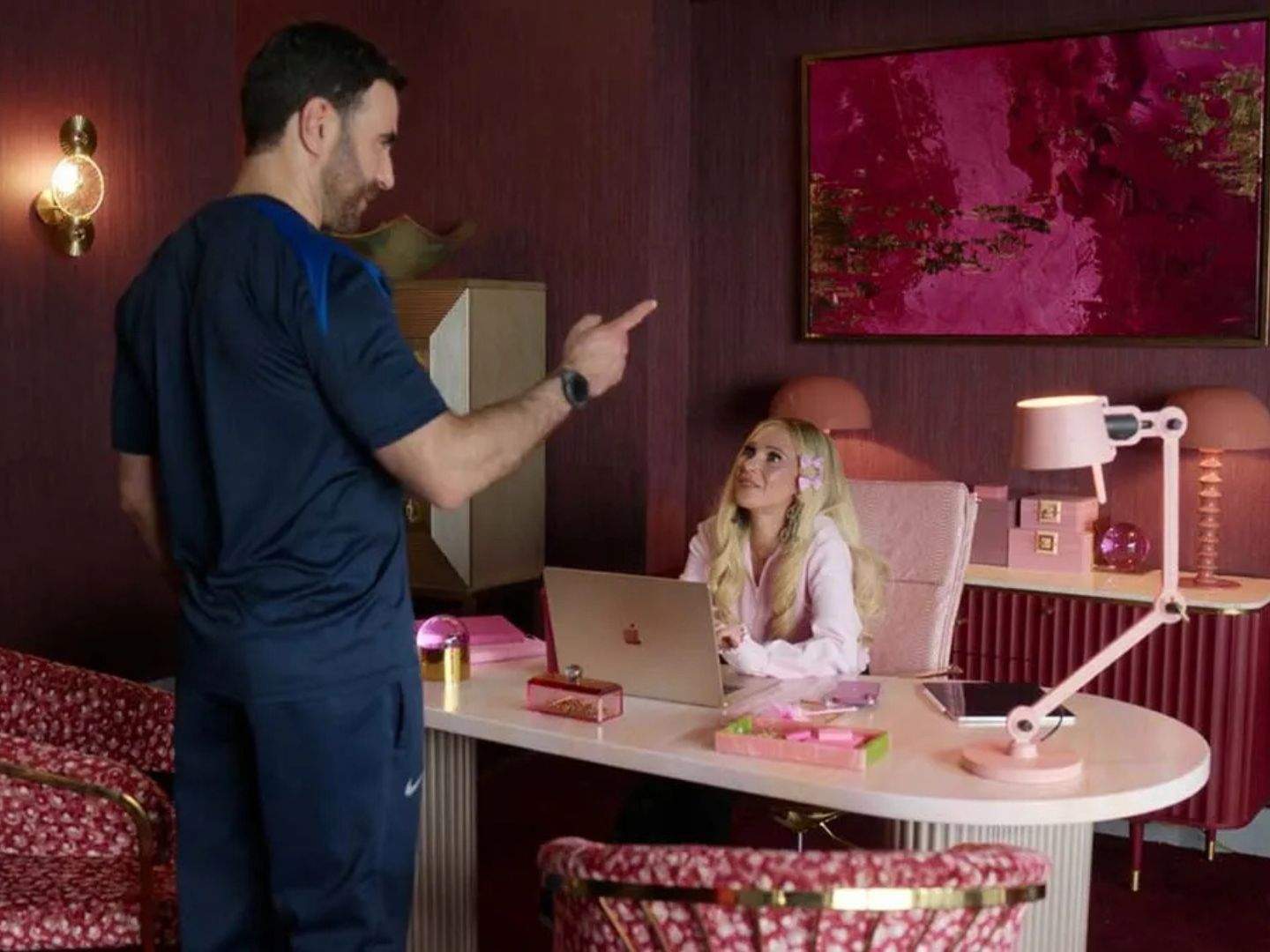 Keeley (Juno Temple), sitting down by her desk, meets Coach Roy (Brett Goldstein), who is standing in front of her, in her pink-themed office.