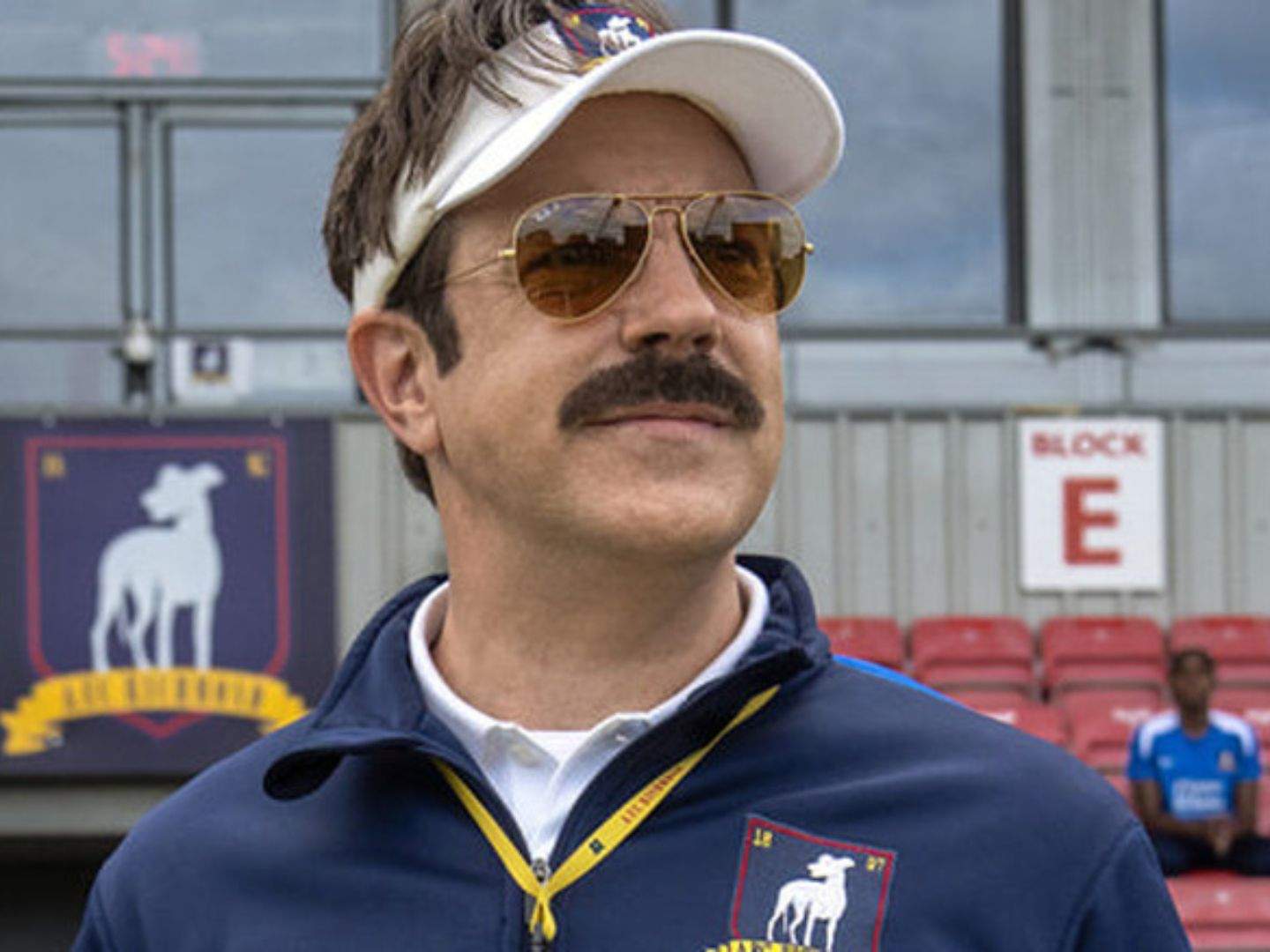 Ted Lasso (Jason Sudeikis), wearing a cap, sunglasses, and a closed jacket, watching the Richmond women's team (not pictured) train.