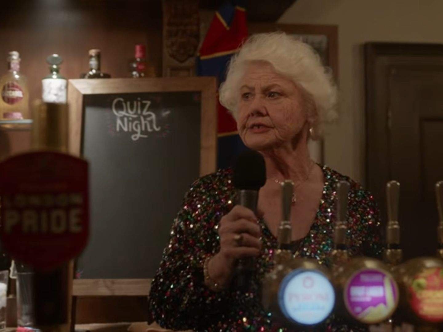 Mae (Annette Badland), the owner of the Crown & Anchor pub, holds a microphone while speaking during quiz night. She wears a sparkling sequined top and stands behind the bar, with a chalkboard reading "Quiz Night" visible in the background.