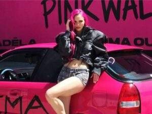 Pink-haired Adéla poses in front of a bright pink hatchback wrapped with the title PRIMA, wearing a black cropped jacket, denim shorts, and black heels. A large pink billboard behind the car promotes her album with the words "Pink Wall."