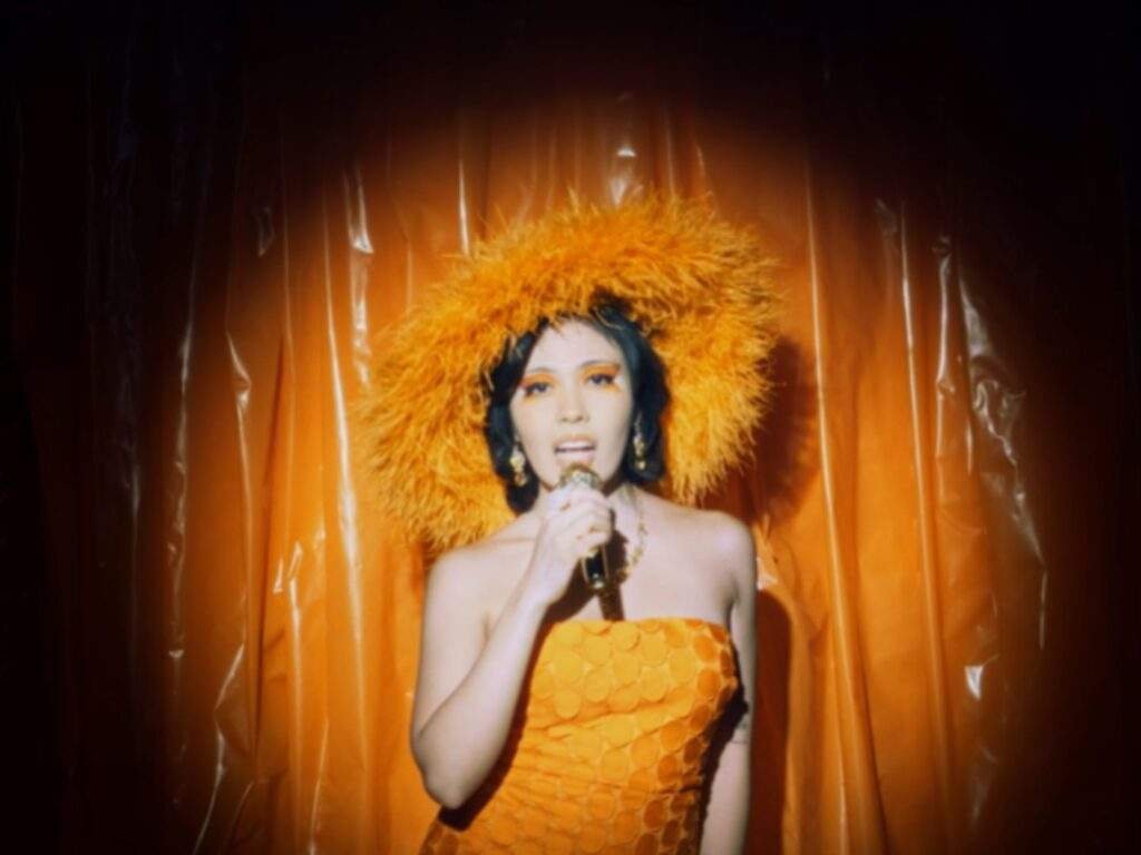 Singer-songwriter kakie performs onstage in a strapless orange dress and a large feathered headpiece, holding a microphone under a spotlight. Warm orange curtains and matching makeup create a bold, theatrical look that complements the performance.