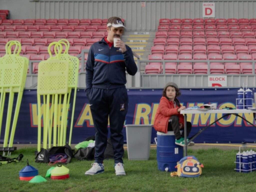 Ted Lasso (Jason Sudeikis) sips coffee while watching Richmond women's team training from the sideline. Beside him, the young son of one of the players sits on a water cooler near the equipment table, with training cones, mannequin dummies, and empty stadium seats in the background.