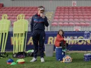 Ted Lasso (Jason Sudeikis) sips coffee while watching Richmond women's team training from the sideline. Beside him, the young son of one of the players sits on a water cooler near the equipment table, with training cones, mannequin dummies, and empty stadium seats in the background.