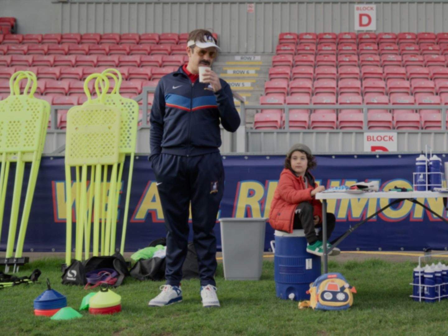 Ted Lasso (Jason Sudeikis) sips coffee while watching Richmond women's team training from the sideline. Beside him, the young son of one of the players sits on a water cooler near the equipment table, with training cones, mannequin dummies, and empty stadium seats in the background.