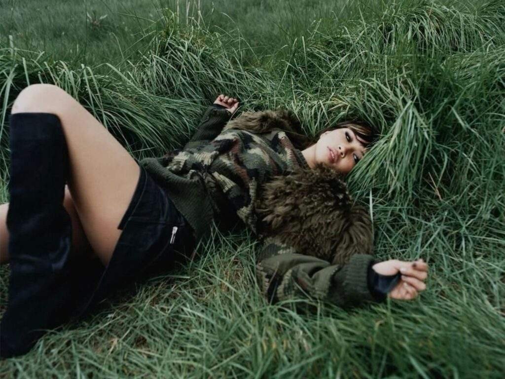 Beabadoobee lies on her back in a patch of tall green grass, looking toward the camera. She wears a camouflage knit sweater with faux fur shoulders, a black mini skirt, and knee-high black boots, creating a moody outdoor portrait with natural greenery filling the background.