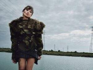 Beabadoobee poses beside a calm body of water under an overcast sky, wearing an oversized camouflage knit sweater with a faux fur collar and a black mini skirt. Power lines and transmission towers stretch across the landscape behind her, creating a stark, industrial backdrop.