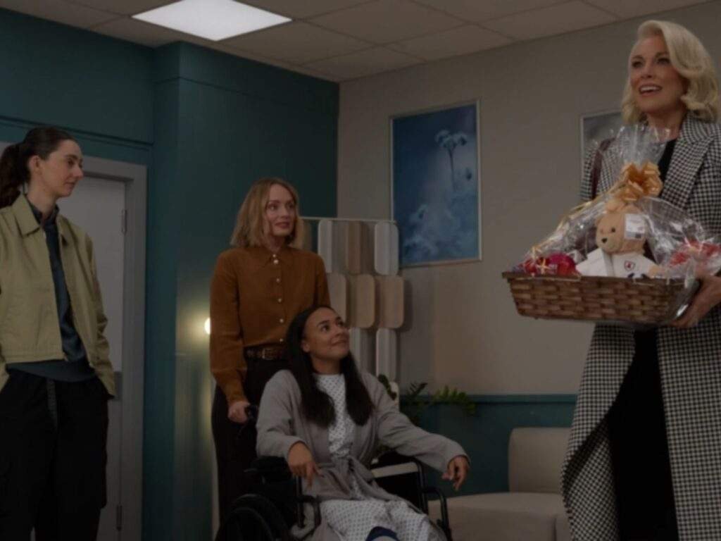 From left to right, Coach Alice Chilton, Dr. Erin Beaumont, Gemma Jay, and Rebecca Welton stand together in a hospital room. Gemma sits in a wheelchair while Rebecca smiles and carries a large gift basket, as Alice and Erin look on. Ted Lasso