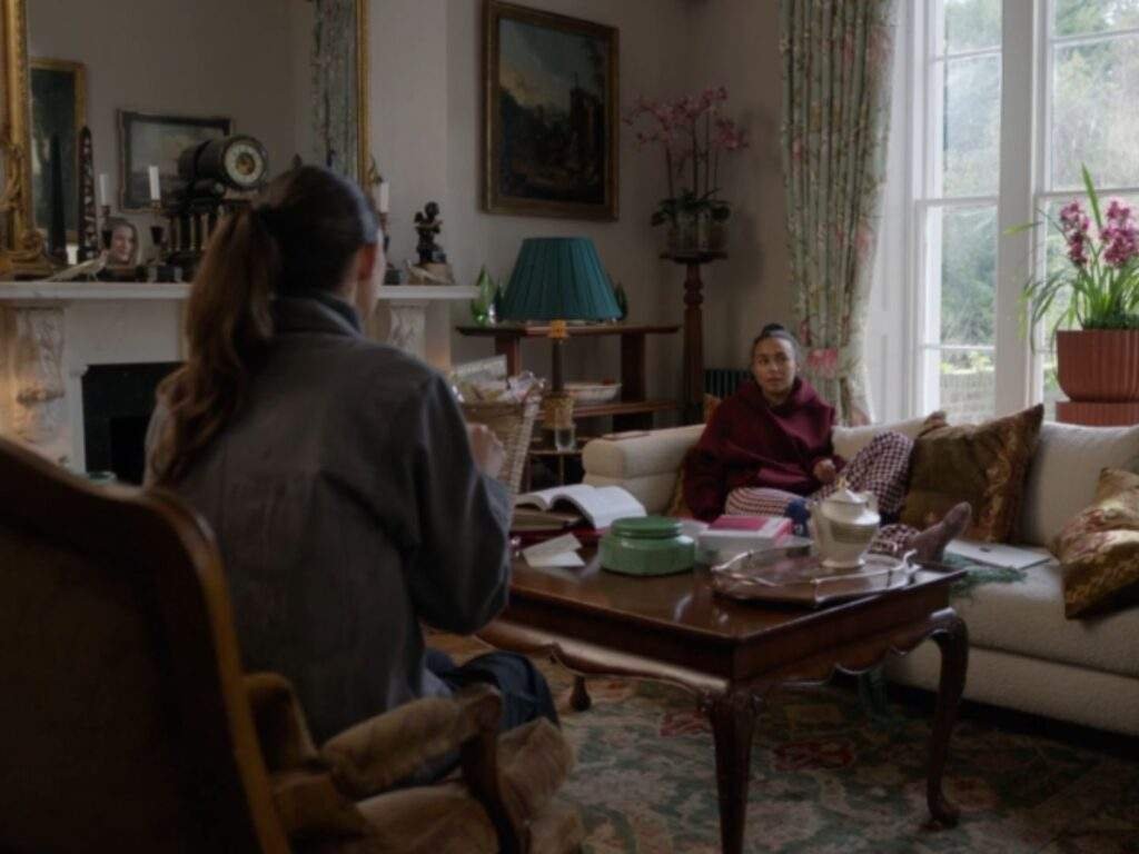 Coach Alice Chilton (Tanya Reynolds) sits facing Gemma Jay (Abbie Hern) during a quiet conversation in a warmly lit living room. Gemma, wearing a maroon hoodie and checkered pajama pants, sits on a sofa beside books and a gift basket as sunlight streams through the large windows.