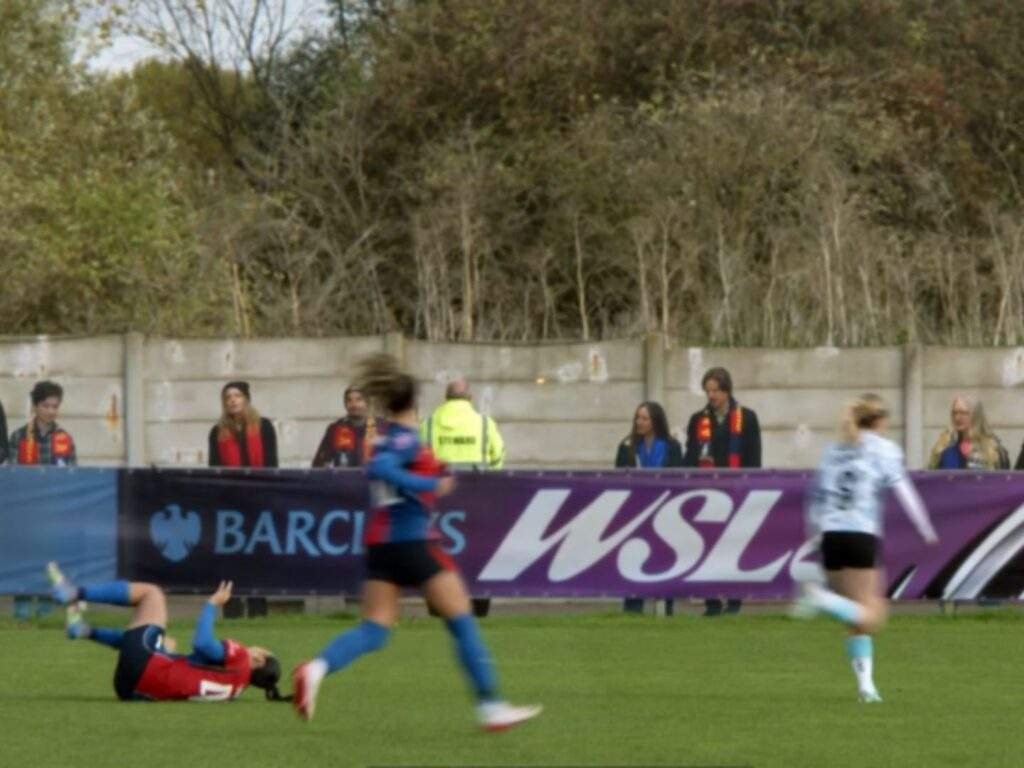 Gemma Jay lies on the pitch after falling during play as nearby players continue chasing the ball. Spectators watch from behind the touchline barriers, with Women's Super League branding visible along the sideline.