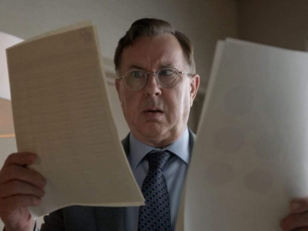 Leslie Higgins (Jeremy Swift) holds two sheets of paper and compares them with a surprised, puzzled expression. Dressed in a gray suit and blue patterned tie, he studies the documents closely while standing indoors.