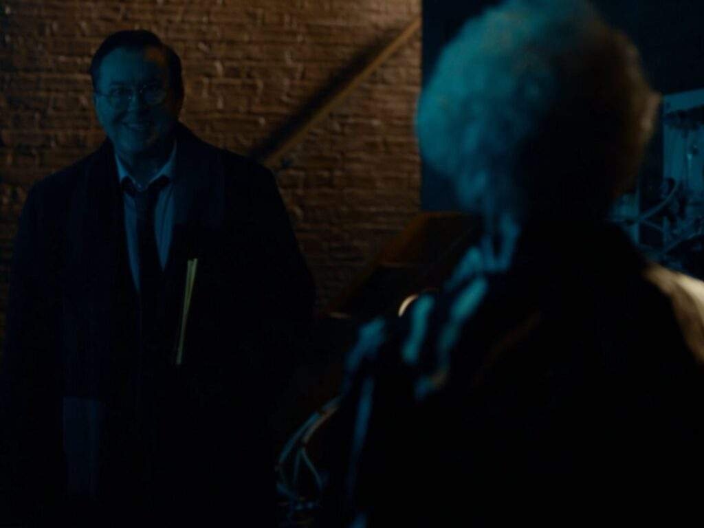 Leslie Higgins (Jeremy Swift) smiles as he speaks with Mae (Annette Badland) in the dimly lit cellar beneath the pub. Higgins stands holding a folder while Mae, seen from behind, faces him amid the shadowy brick-lined room.