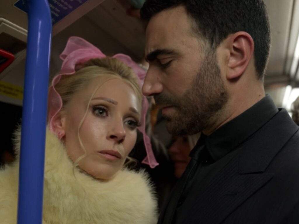 Keeley Jones and Roy Kent stand closely together inside a crowded train carriage. Keeley, wearing a white faux fur coat and a large pink bow in her hair, looks up at Roy with a serious, emotional expression while Roy, dressed in a dark suit, looks down pensively as the two share a quiet, tense moment. Ted Lasso