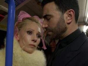 Keeley Jones and Roy Kent stand closely together inside a crowded train carriage. Keeley, wearing a white faux fur coat and a large pink bow in her hair, looks up at Roy with a serious, emotional expression while Roy, dressed in a dark suit, looks down pensively as the two share a quiet, tense moment. Ted Lasso