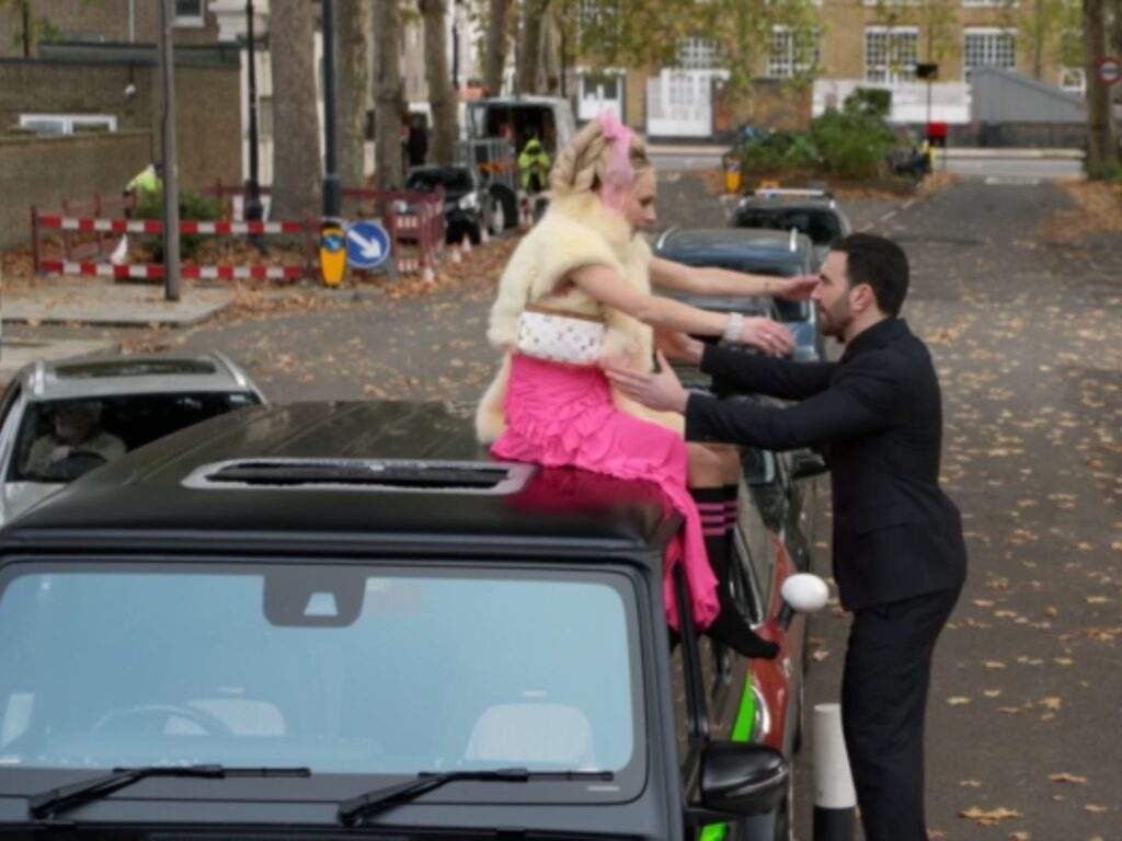 Keeley Jones reaches out to embrace Roy Kent as she sits on the roof of a parked car along a quiet, tree-lined street. Roy stands beside the vehicle with his arms open while autumn leaves scatter across the road behind them.