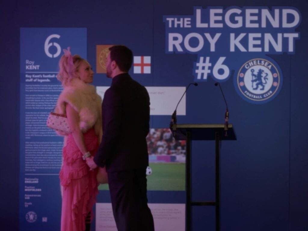 Keeley Jones and Roy Kent stand face to face, holding hands during a quiet, emotional moment. Behind them, a museum-style display honoring Roy's football career, including a large sign reading "The Legend Roy Kent #6," forms the backdrop.