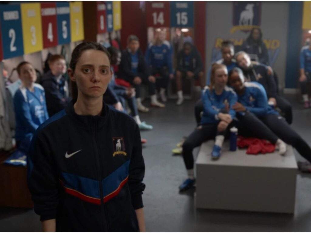 Coach Alice Chilton  (Tanya Reynolds) stands at the front of the Lady Greyhounds' locker room with a serious, thoughtful expression. The players sit and stand quietly in the background, watching. Ted Lasso