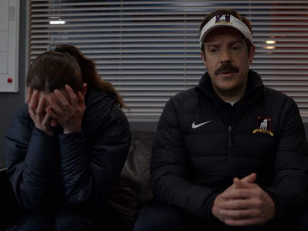 Coach Alice Chilton sits with her face buried in her hands as Ted Lasso sits beside her, speaking calmly in the team office. Ted looks toward Alice with a concerned expression while she appears emotionally overwhelmed after a difficult moment.
