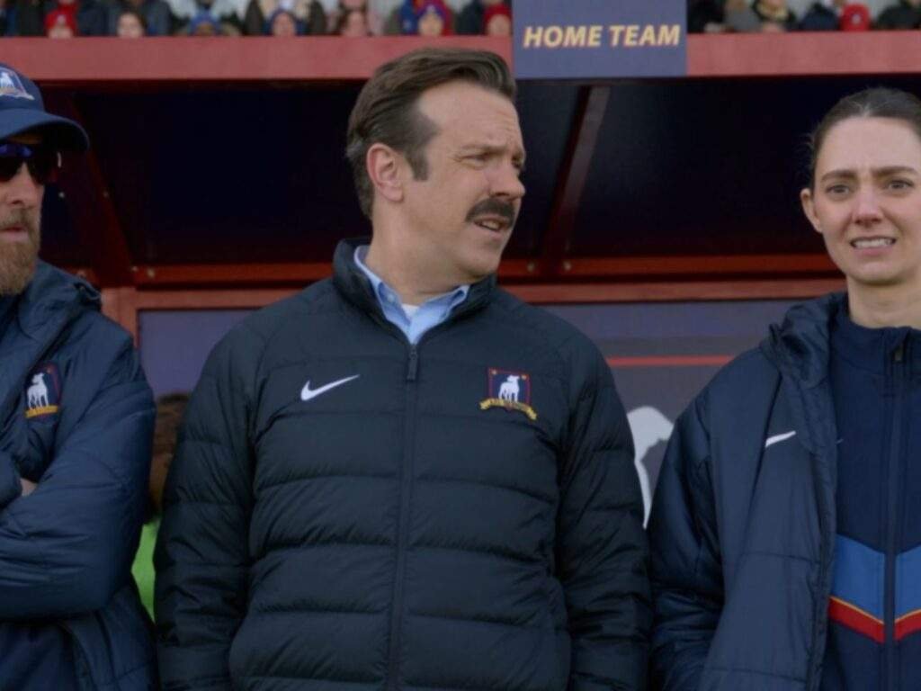 Coach Beard, Ted Lasso, and Coach Alice Chilton stand together on the touchline during a match. Ted, in the center, looks across the field with a concerned expression while Beard and Alice watch from either side, with the team bench and stadium crowd visible in the background.