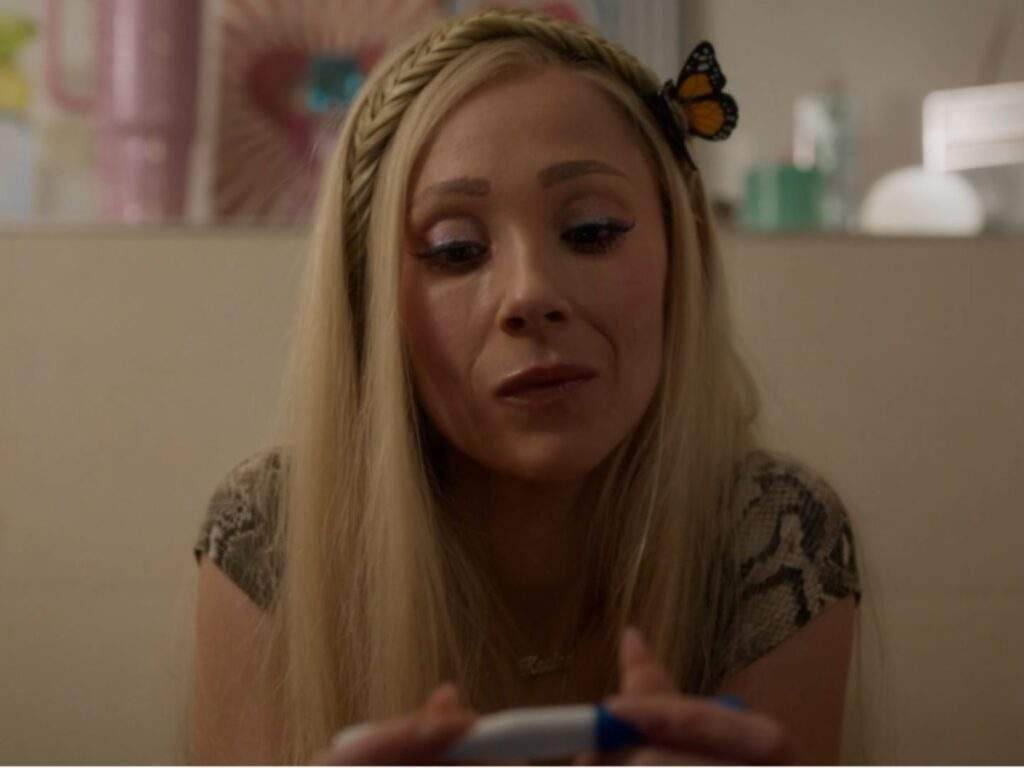 Keeley Jones (Juno Temple) looks down with a thoughtful, emotional expression while holding a pregnancy test, with a butterfly hair clip in her hair and a softly lit room in the background.