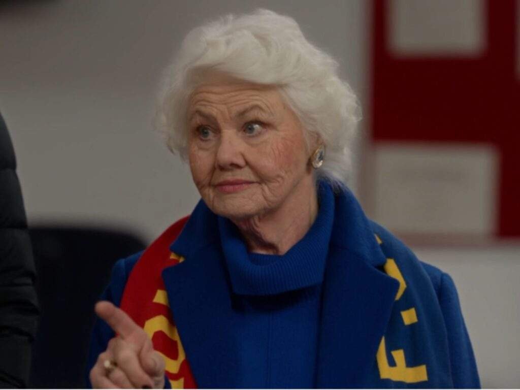 Mae Green (Annette Badland) points forward with a warm but firm expression while wearing a blue outfit and Richmond scarf, standing indoors during a conversation.