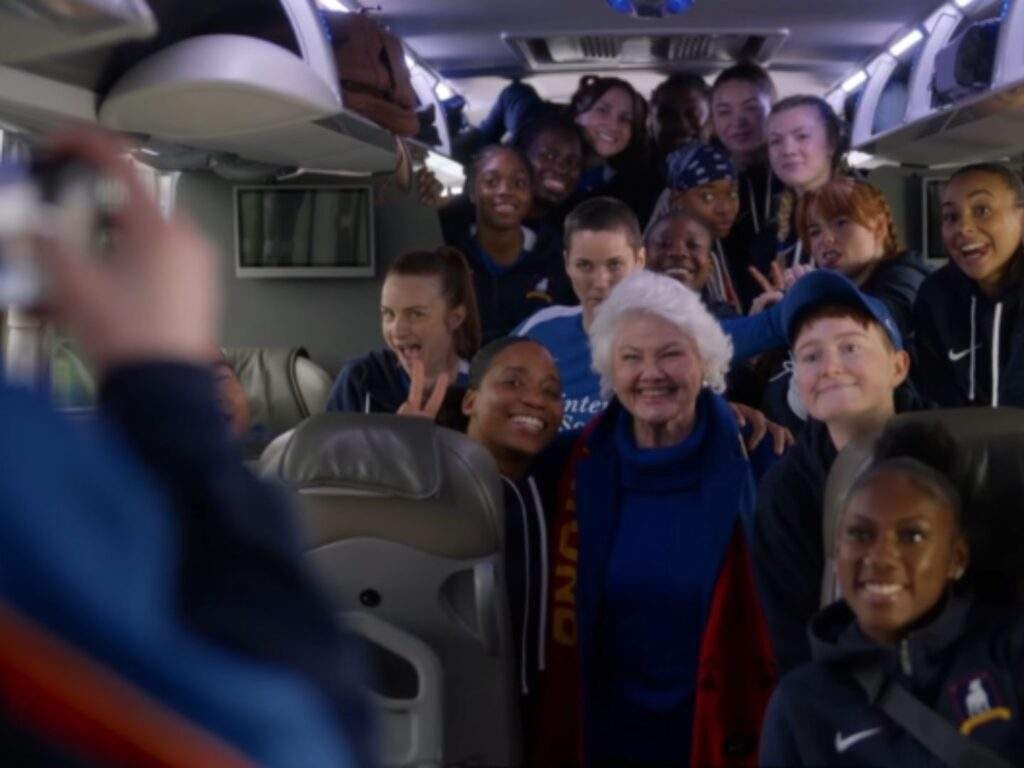 The AFC Richmond's women's team gather inside the team bus for a group photo with Mae Green (Annette Badland), who smiles at the center wearing a Richmond scarf. Coach Alice Chilton (Tanya Reynolds), not pictured, takes the photo from the front of the bus with her phone. Ted Lasso season 4