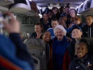The AFC Richmond's women's team gather inside the team bus for a group photo with Mae Green (Annette Badland), who smiles at the center wearing a Richmond scarf. Coach Alice Chilton (Tanya Reynolds), not pictured, takes the photo from the front of the bus with her phone. Ted Lasso season 4