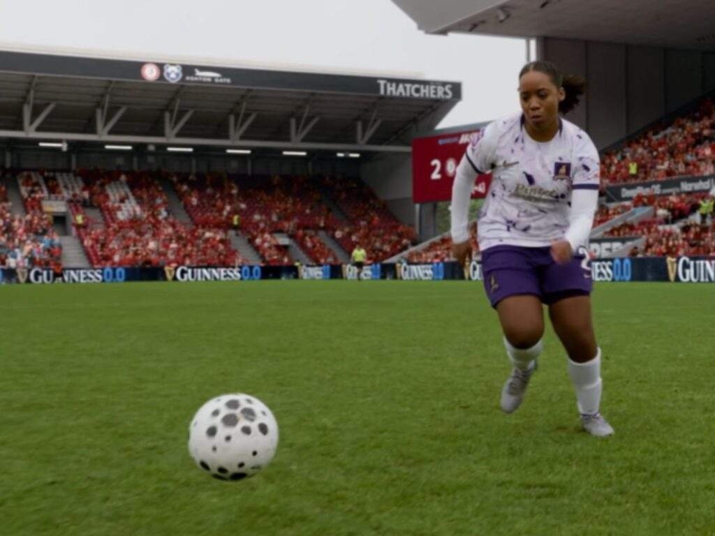 Shannon (Shannon Hayes) runs toward the football during a match for the Lady Greyhounds, with the stadium crowd and scoreboard visible in the background as she closes in on the ball. Ted Lasso season 4