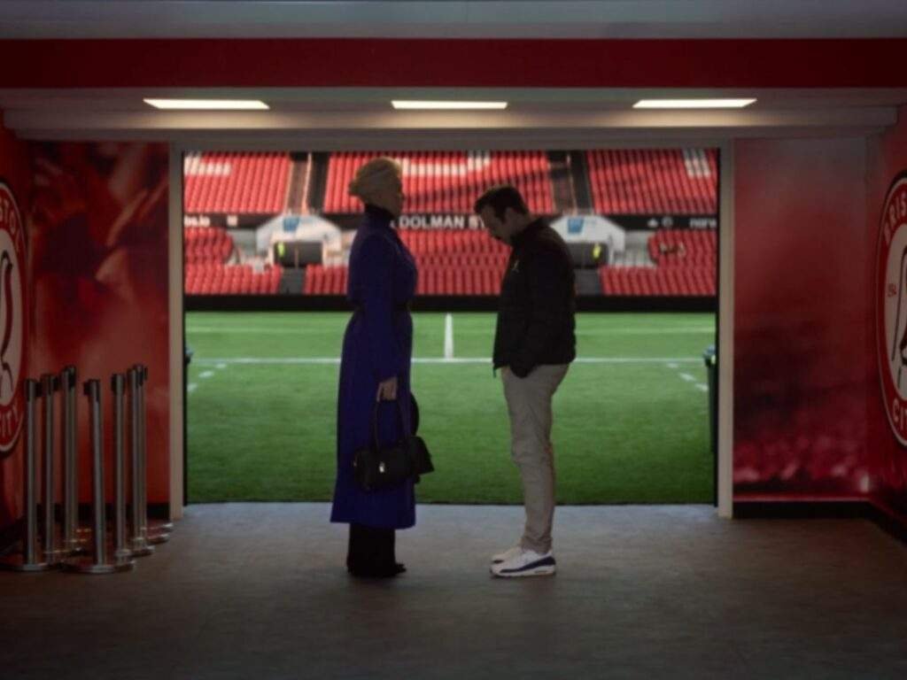 Rebecca Welton (Hannah Waddingham) stands with Ted Lasso (Jason Sudeikis) in the stadium tunnel as she shares the news of Mae Green's sudden passing. Ted looks down in stunned silence while Rebecca faces him, with the empty pitch visible behind them. Ted Lasso season 4