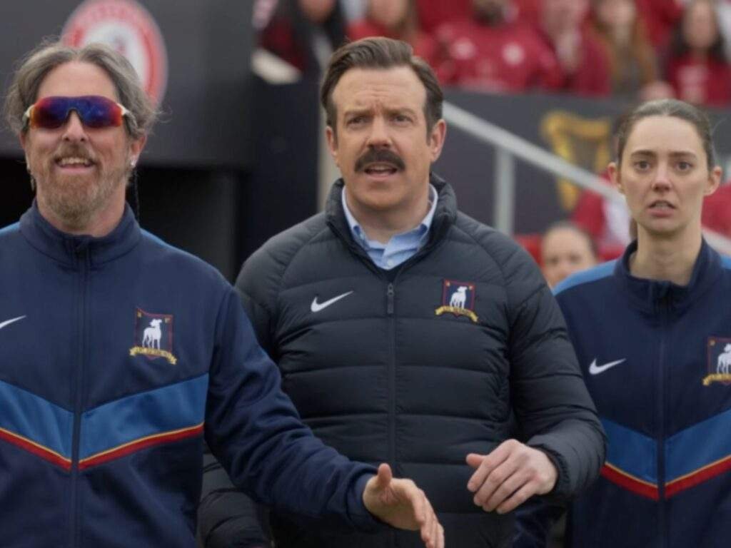 Coach Beard (Brendan Hunt), Ted Lasso (Jason Sudeikis), and Coach Alice Chilton (Tanya Reynolds) stand together on the touchline during a match, watching the action with tense expressions. Beard gestures with one hand while Ted and Alice focus intently on the field.