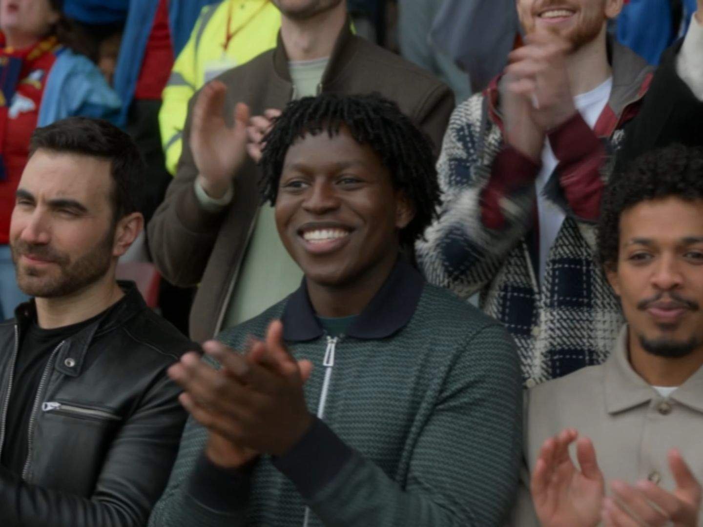 Roy Kent (Brett Goldstein) and Sam Obisanya (Toheeb Jimoh) applaud from the stands as they watch the Lady Greyhounds play, joined by other AFC Richmond supporters cheering around them.