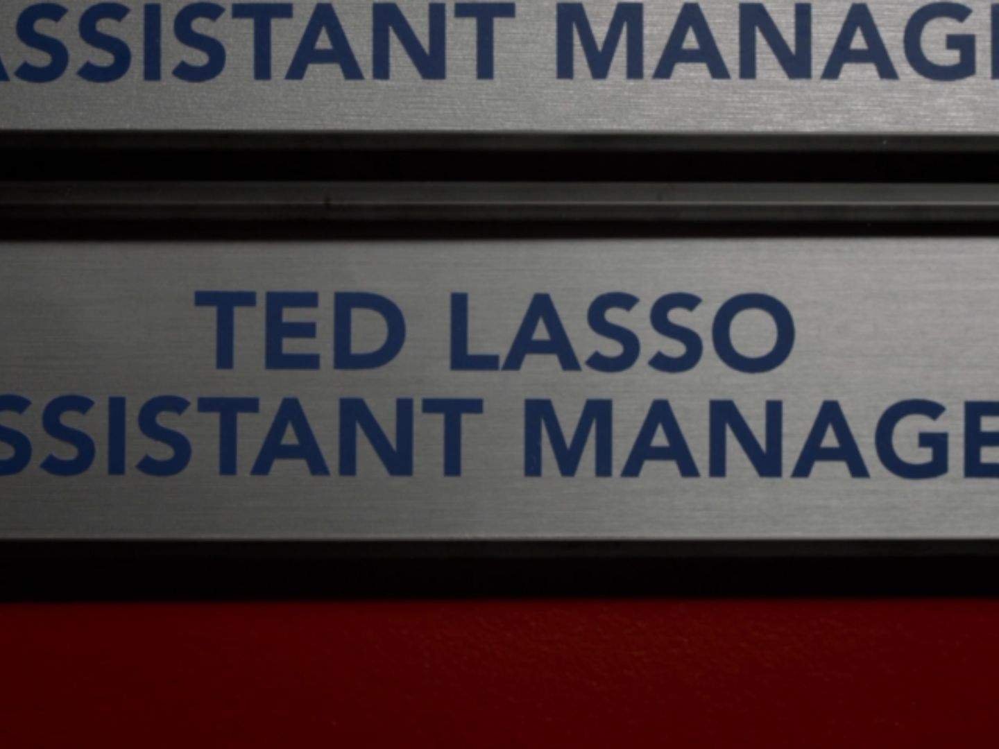 Alt text: A close-up of a nameplate on an office door identifies Ted Lasso as the assistant manager of the AFC Richmond women's team.