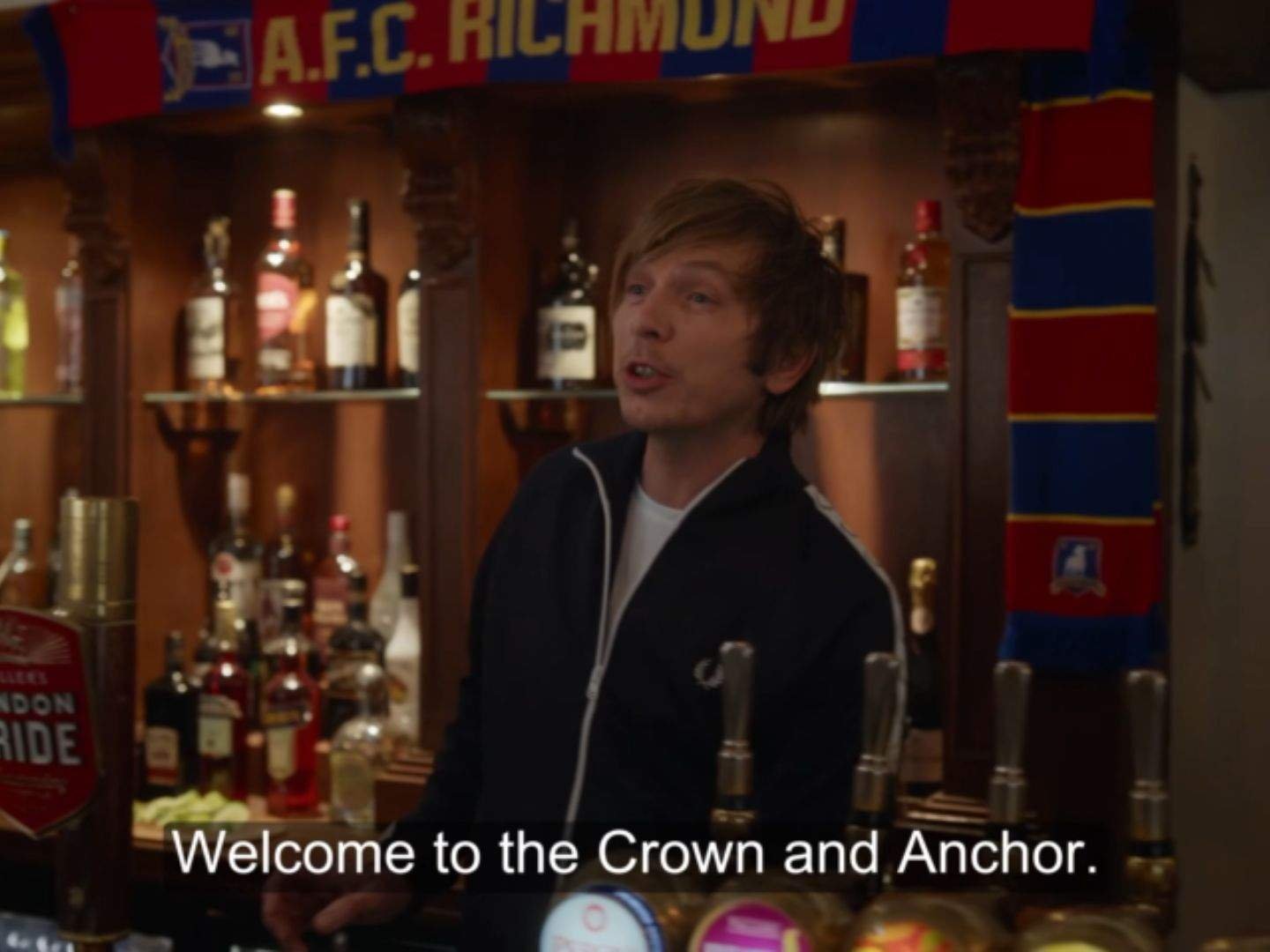 Baz (Adam Colborne) stands behind the bar at the Crown & Anchor pub, welcoming customers beneath an AFC Richmond scarf with shelves of bottles behind him.