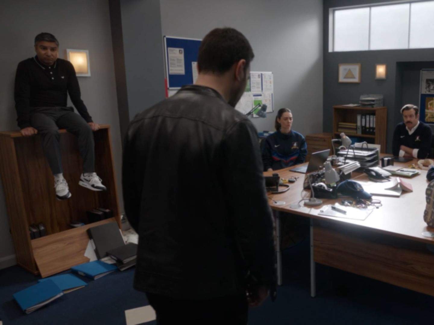 Roy Kent (Brett Goldstein) stands in the center of the AFC Richmond coaches' office during a Diamond Dogs meeting as Nate Shelley (Nick Mohammed) sits on a cabinet, Coach Alice Chilton (Tanya Reynolds) sits at the desk, and Ted Lasso (Jason Sudeikis) listens from across the room.