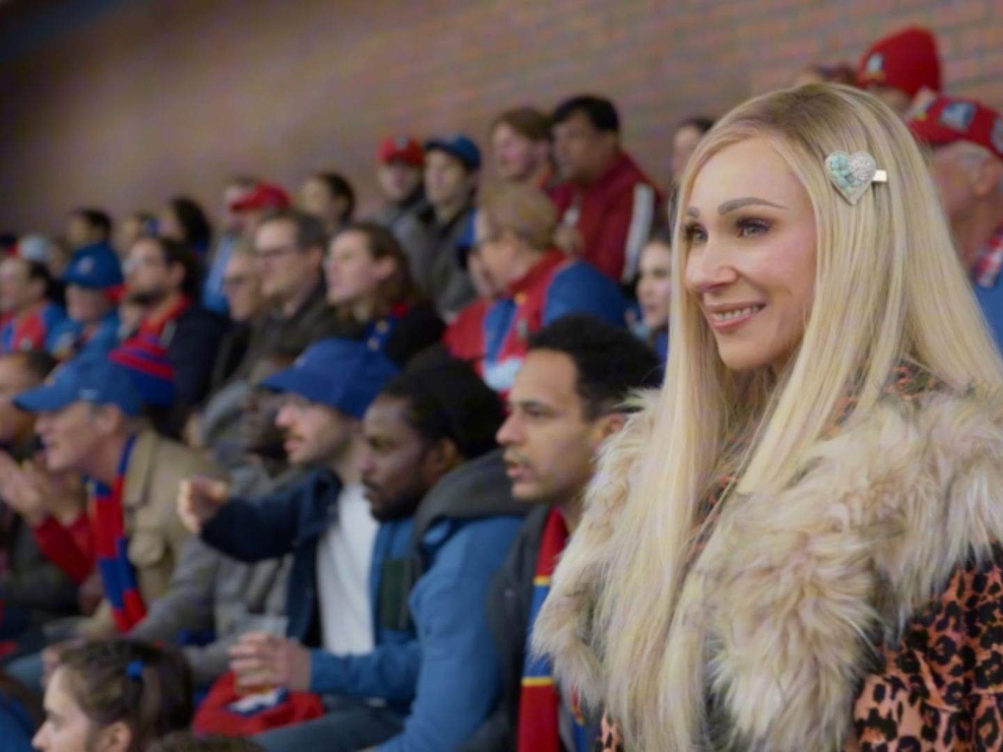 Keeley Jones (Juno Temple) smiles from the stands as she watches the Lady Greyhounds play, wearing a leopard-print coat with a fur collar and a heart-shaped hair clip among AFC Richmond supporters.