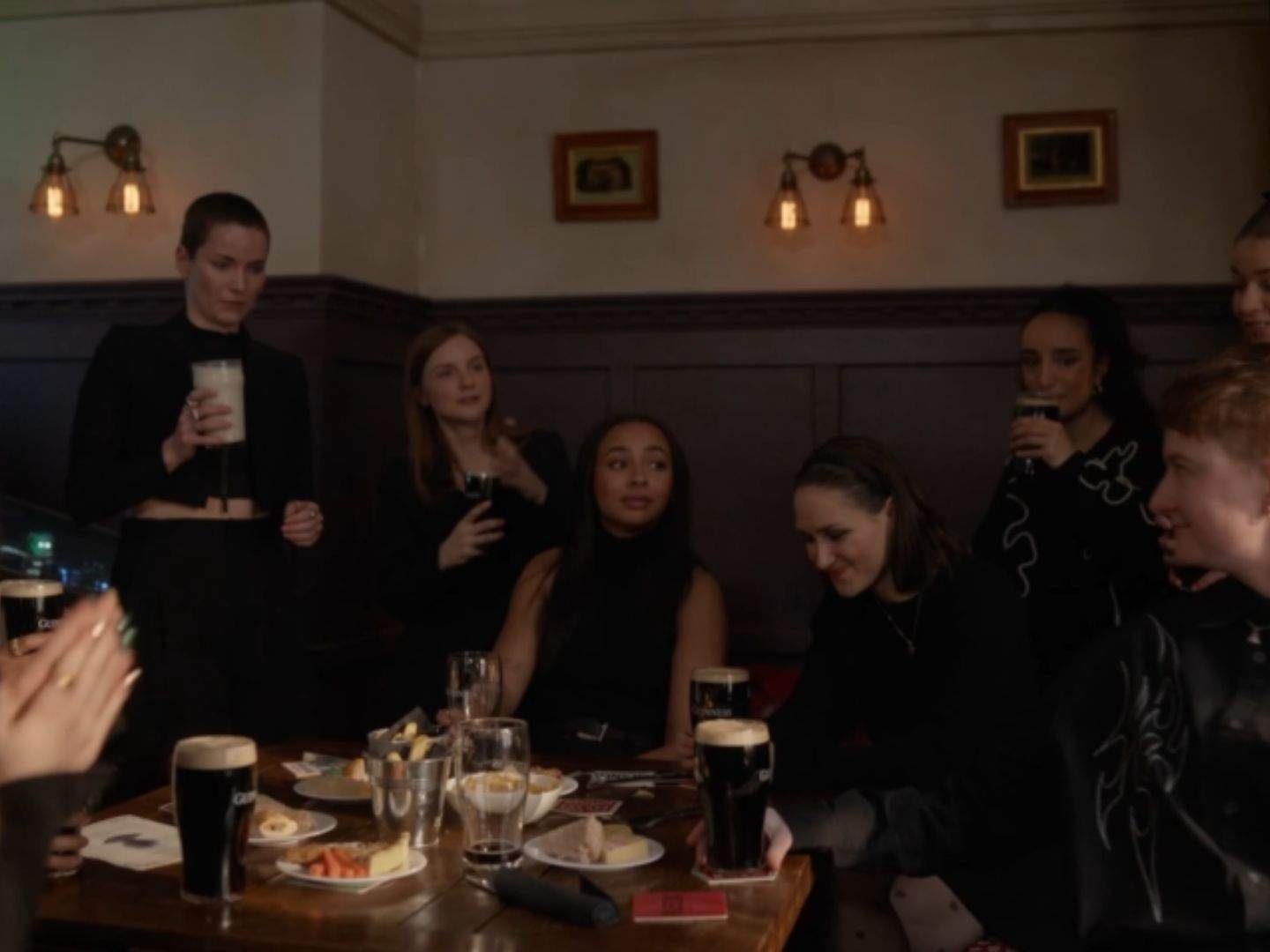 Members of the Lady Greyhounds gather around a table at Mae Green's memorial in the Crown & Anchor, holding pints and talking quietly as food and playing cards sit on the table.