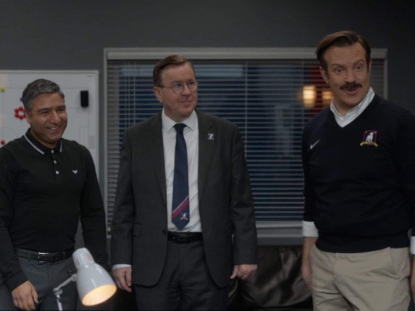 Nate Shelley (Nick Mohammed), Leslie Higgins (Jeremy Swift), and Ted Lasso (Jason Sudeikis) stand together in the AFC Richmond coaches' office, smiling as they look toward someone off camera.