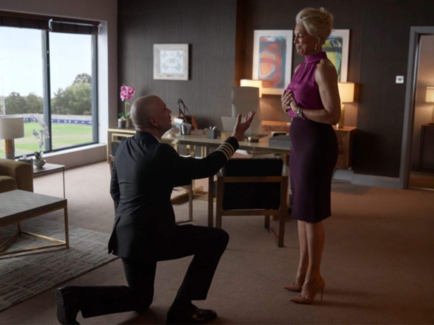 Matthijs (Matteo van der Grijn) kneels on one knee as he proposes to Rebecca Welton (Hannah Waddingham) in her office, while Rebecca stands with one hand over her chest, visibly surprised and emotional.