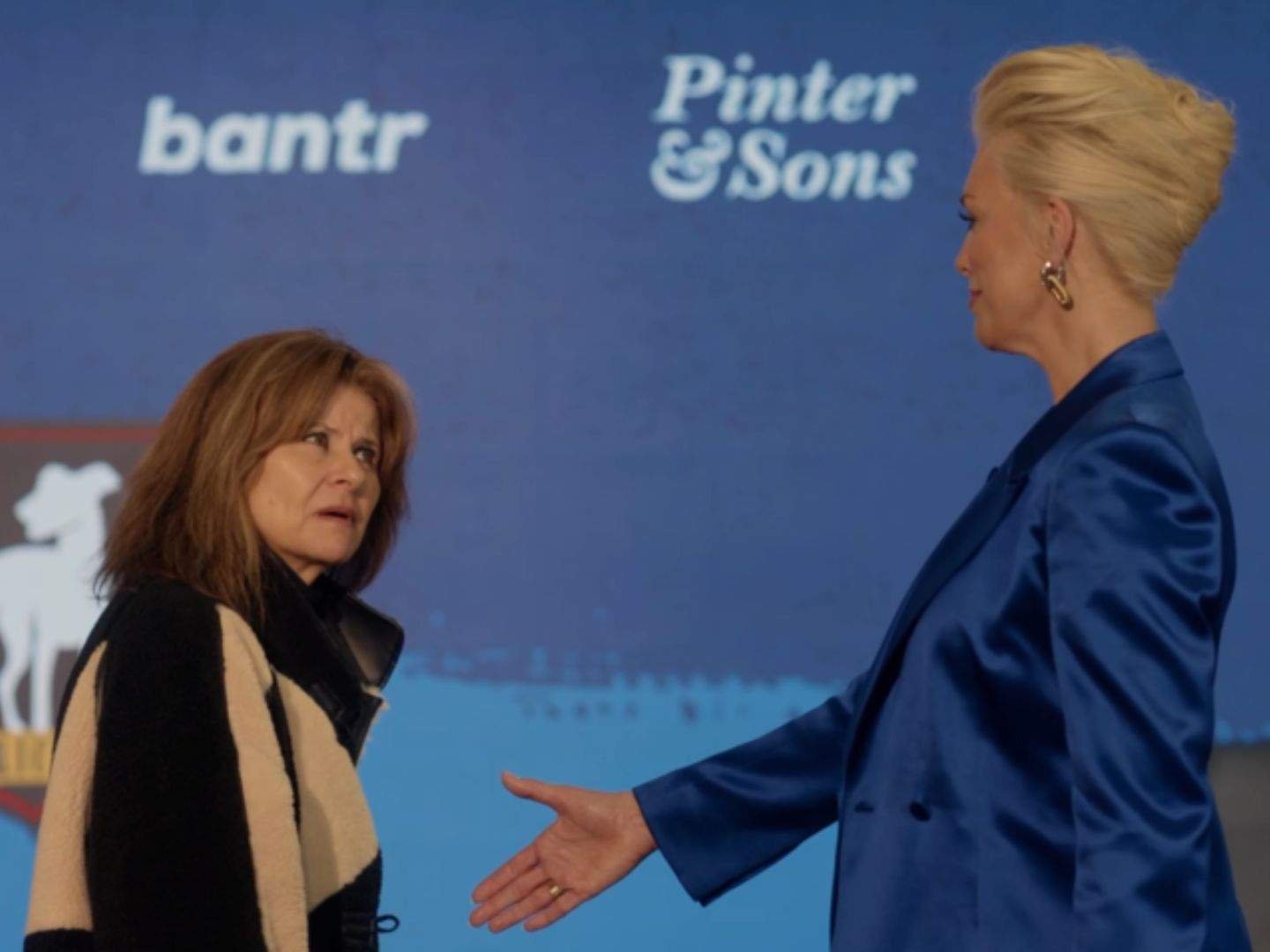 Zelda Southport (Tracey Ullman) faces Rebecca Welton (Hannah Waddingham) and extends her hand for a handshake during a tense encounter in front of a presentation screen displaying AFC Richmond sponsors.