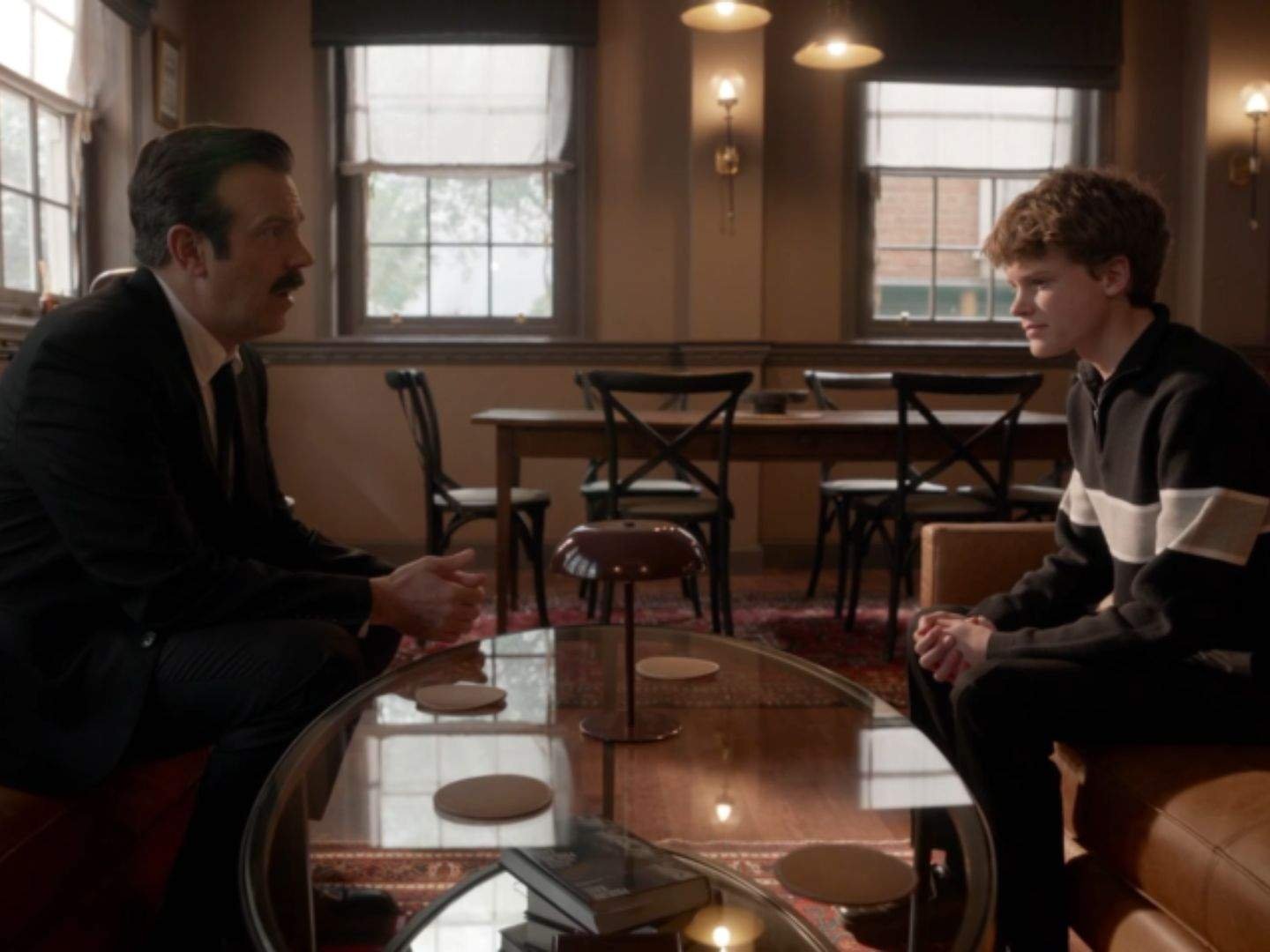 Ted Lasso (Jason Sudeikis) sits across from his son Henry (Grant Feely) in their living room as the two share a serious, heartfelt conversation while facing each other across a coffee table.