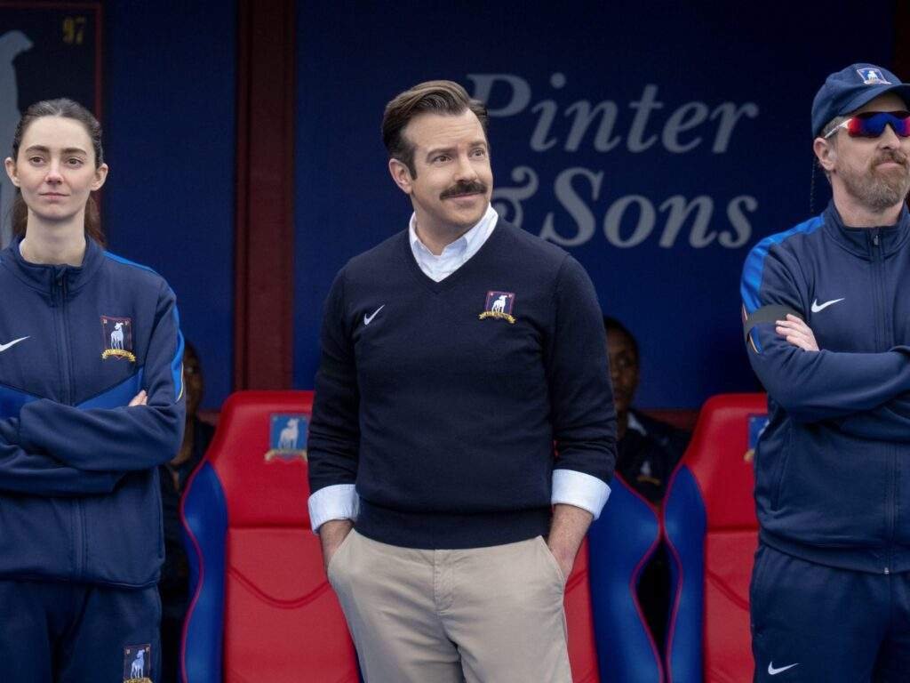Coach Alice Chilton (Tanya Reynolds), Ted Lasso (Jason Sudeikis), and Coach Beard (Brendan Hunt) stand in front of the AFC Richmond dugout during a match, watching the action from the touchline.