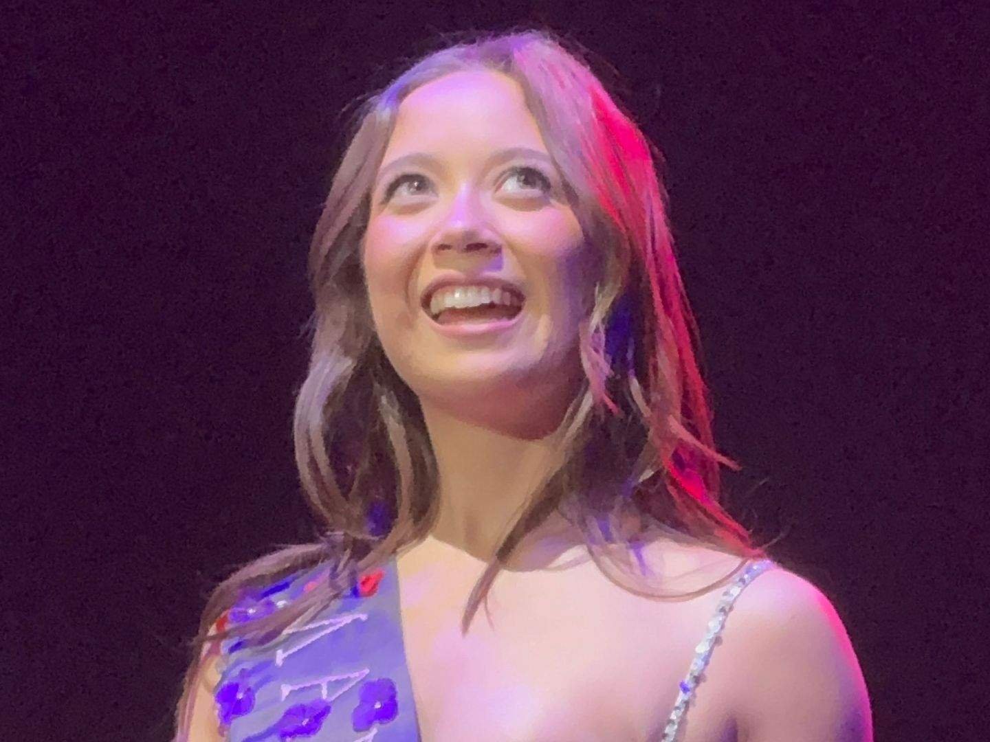 Laufey smiles brightly onstage while looking upward, wearing a sparkling light-colored dress under purple and pink stage lights during a concert in Manila.