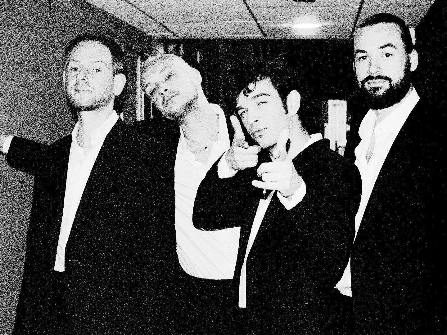 Members of The 1975 pose together backstage in matching black suits and white shirts, with Matty Healy pointing toward the camera in a black-and-white photo.